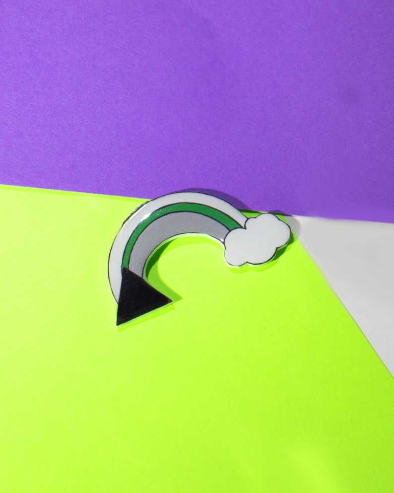 May include: A silver-colored pin shaped like a rainbow with a black triangle at the bottom and a white cloud at the top. The rainbow has green, gray, and white stripes.