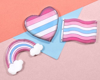May include: Three colorful pins with a transgender pride theme. A pink, blue, and white striped heart, a rainbow-striped rainbow, and a pink, blue, and white striped flag.