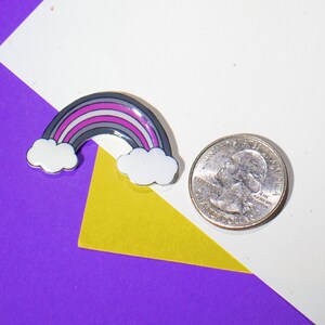 Op de afbeelding: Een kleine regenboogspeld met een grijs, wit, roze en paars ontwerp. De speld ligt op een wit oppervlak met een gele en paarse achtergrond.