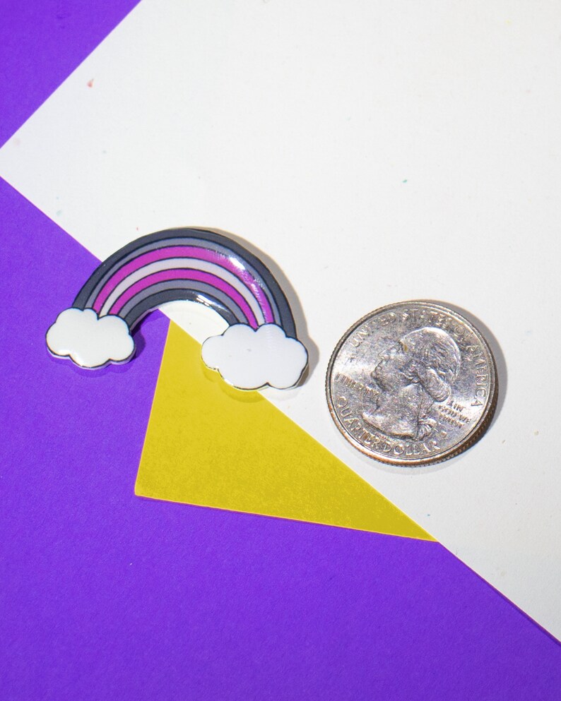 Op de afbeelding: Een kleine regenboogspeld met een grijs, wit, roze en paars ontwerp. De speld ligt op een wit oppervlak met een gele en paarse achtergrond.