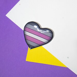 Op de afbeelding: Een hartvormige pin met een gestreept ontwerp in grijs, wit en roze. De pin is op een witte en paarse achtergrond.