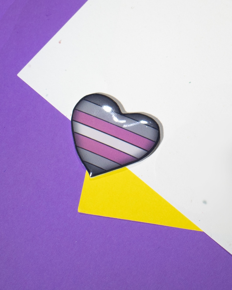 Op de afbeelding: Een hartvormige pin met een gestreept ontwerp in grijs, wit en roze. De pin is op een witte en paarse achtergrond.