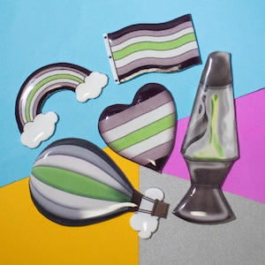 Op de afbeelding: Vijf kleurrijke, decoratieve pinnen met een groen, wit en grijs kleurenschema. De pinnen zijn gevormd als een regenboog, een hart, een vlag, een luchtballon en een lava lamp.