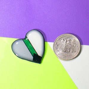 May include: A heart-shaped pin with a green, gray, and black design. The pin is next to a silver US quarter with the inscription "In God We Trust" and "Liberty" on the coin.