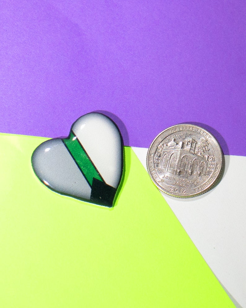 May include: A heart-shaped pin with a green, gray, and black design. The pin is next to a silver US quarter with the inscription "In God We Trust" and "Liberty" on the coin.