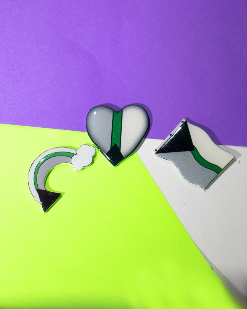 May include: Three colorful pins with a green, white, and black color scheme. The pins are shaped like a heart, a rainbow, and a flag. The pins are on a purple, white, and green background.