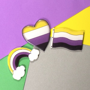 May include: Three yellow, white and purple pins in the shape of a heart, a rainbow and a flag. The pins are on a purple, yellow and grey background.