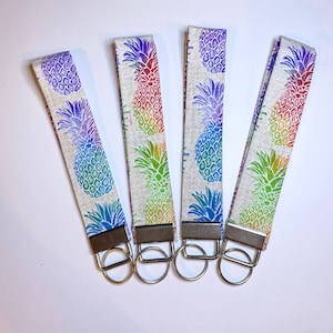 May include: Four key fobs with a repeating rainbow pineapple pattern on a light gray background. Each fob has a silver-colored metal clasp and key ring. The pineapple design transitions from purple to green.