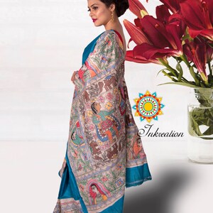 May include: A woman wearing a teal blue saree with a white and colorful hand-painted design. The saree features a traditional Indian folk art style with figures and animals. The saree is draped over her shoulder and waist, and she is standing in front of a white background.