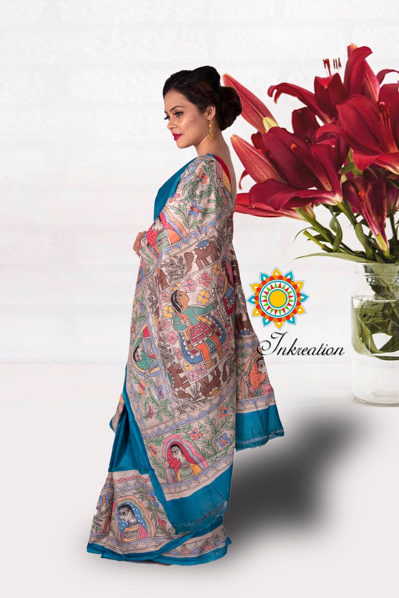 May include: A woman wearing a teal blue saree with a white and colorful hand-painted design. The saree features a traditional Indian folk art style with figures and animals. The saree is draped over her shoulder and waist, and she is standing in front of a white background.