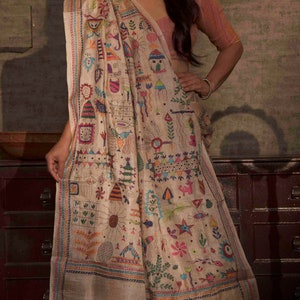 May include: A woman wearing a cream-coloured saree with colourful embroidery. The saree features a variety of patterns, including flowers, birds, and geometric shapes. The saree is draped over a pink blouse. The woman is standing in front of a brown wall.