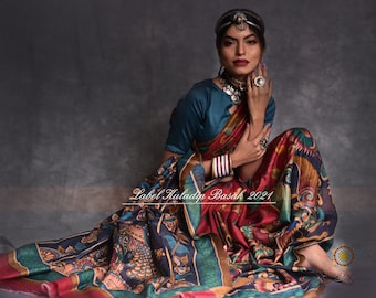 May include: A woman wearing a teal blue blouse and a colorful patterned saree with gold accents. She is wearing a silver headpiece and jewelry. The saree has a red, blue, green, and gold color scheme. The text "Label Khuladip Basak 2021" is visible in the image.