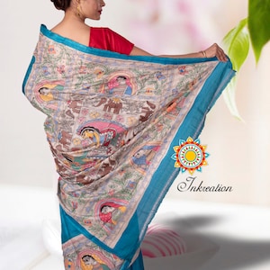 May include: A woman wearing a blue and cream saree with a colorful hand-painted design featuring people and animals. The saree has a blue border and a cream base with intricate details. The design is reminiscent of traditional Indian art.
