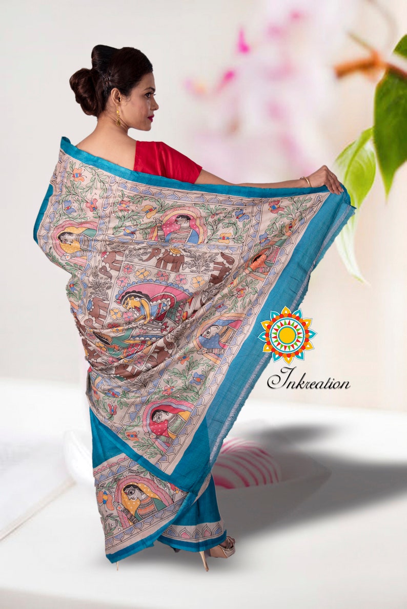May include: A woman wearing a blue and cream saree with a colorful hand-painted design featuring people and animals. The saree has a blue border and a cream base with intricate details. The design is reminiscent of traditional Indian art.