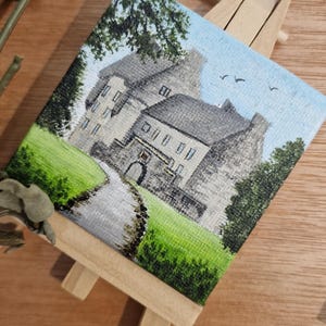 Può includere: Piccolo dipinto di un castello in pietra su un cavalletto di legno. L'opera raffigura un sentiero che conduce al castello, circondato da erba verde e alberi, sotto un cielo blu con uccelli. È un'opera d'arte in miniatura.