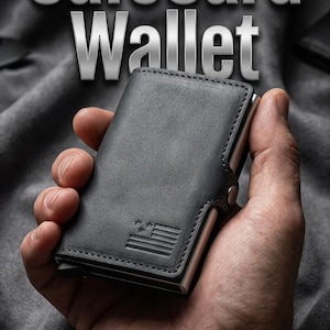 May include: A gray SafeCard Wallet with a metal frame is held in a hand. The wallet has a small American flag design. The words "SafeCard Wallet" are displayed in a silver, metallic font above the wallet.