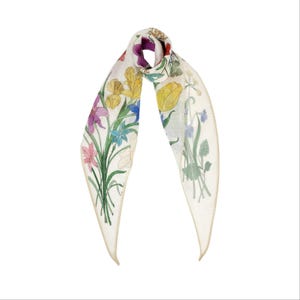 May include: A white scarf with a floral design featuring yellow, purple, pink, and blue flowers. The scarf is rectangular with a gold trim and is tied in a knot.
