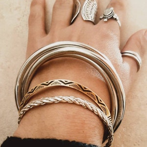 May include: A hand adorned with silver rings and bracelets. The bracelets include thick, smooth bands, a gold band with a black geometric design, and a braided silver bracelet. The rings feature intricate designs.