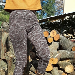 May include: Brown leggings with a white geometric pattern. The leggings are being worn by a person standing on a pile of logs.