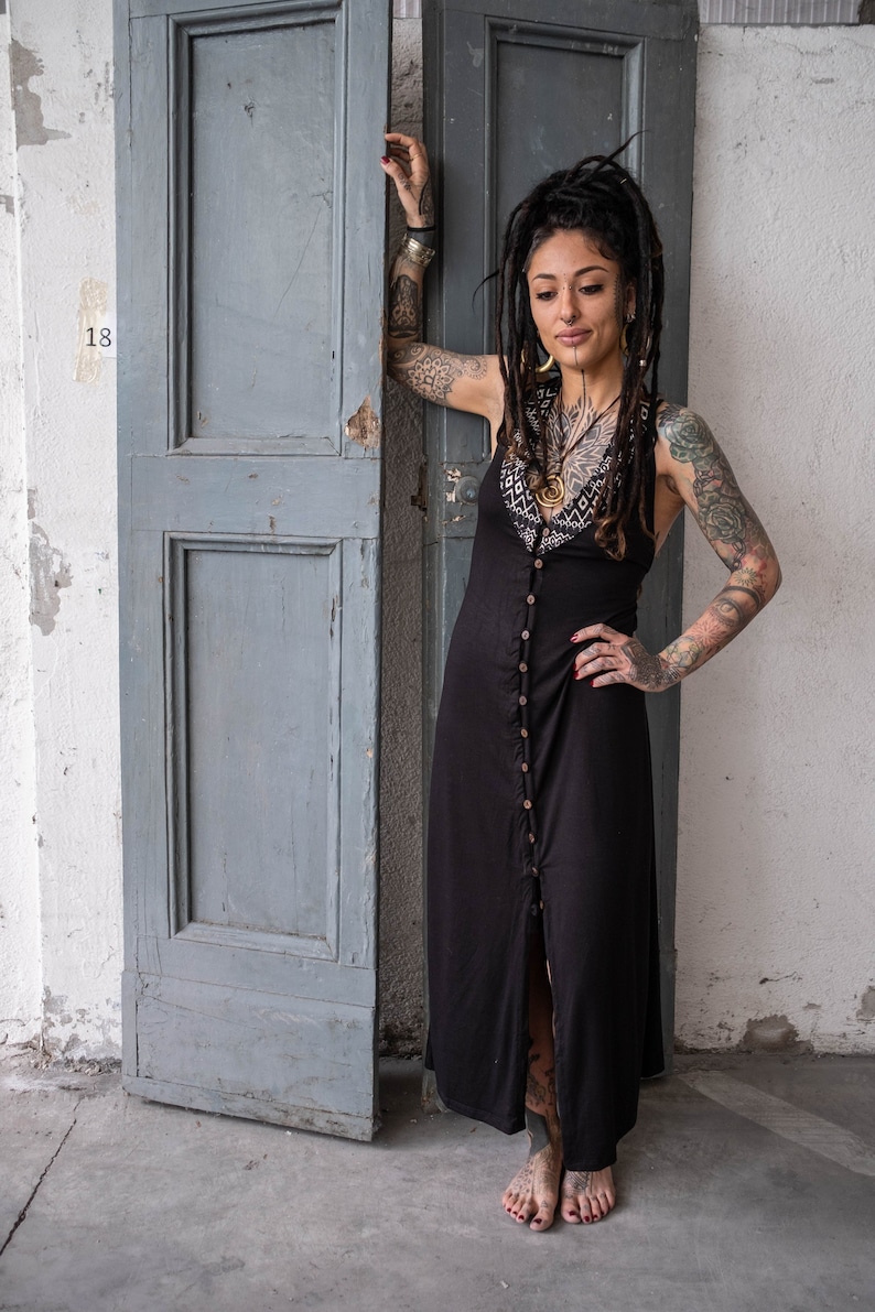 May include: Black sleeveless maxi dress with a button-down front and a decorative patterned yoke. The dress is worn by a person with tattoos and dreadlocks, standing in front of a weathered gray door.