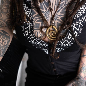 May include: Black top with a white geometric pattern along the neckline. A gold spiral pendant hangs from a brown cord necklace. The person has multiple tattoos on their arms and chest, and is wearing bracelets.