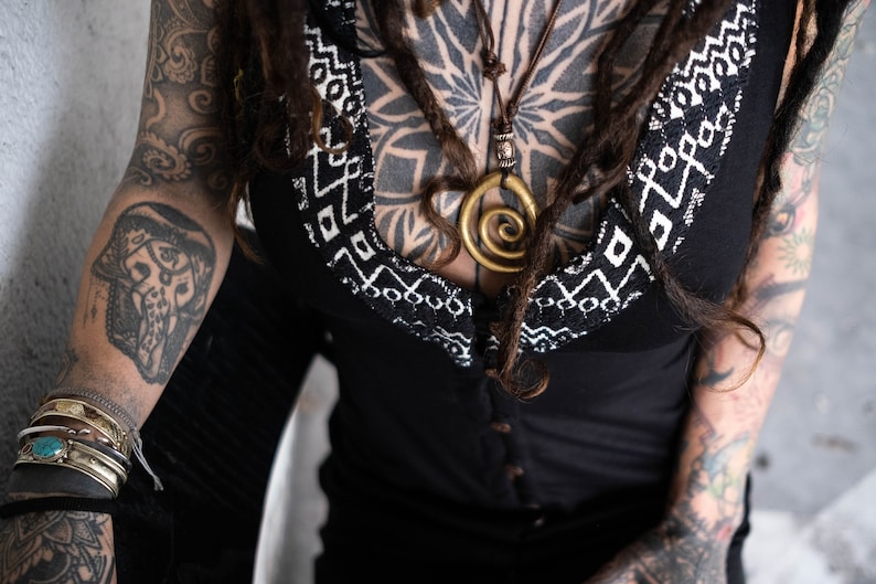 May include: Black top with a white geometric pattern along the neckline. A gold spiral pendant hangs from a brown cord necklace. The person has multiple tattoos on their arms and chest, and is wearing bracelets.
