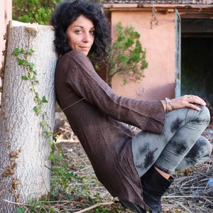 May include: A person wearing a long-sleeved brown tunic and grey tie-dye leggings, leaning against a tree trunk. She is wearing black boots and silver jewellery. The background includes a pink building and a blue door.