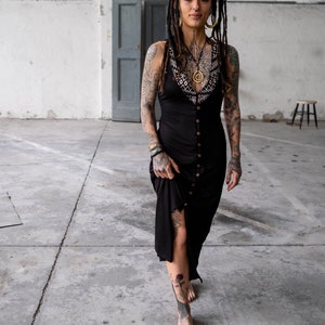 May include: A black button-down dress with a decorative neckline. The dress is knee-length and has a slit on the side. The person in the image has tattoos on their arms, chest, and legs. They are wearing gold earrings and a necklace.