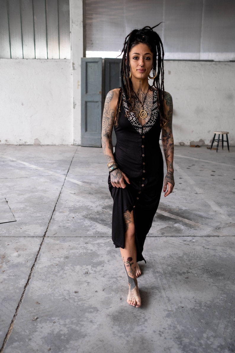 May include: A black button-down dress with a decorative neckline. The dress is knee-length and has a slit on the side. The person in the image has tattoos on their arms, chest, and legs. They are wearing gold earrings and a necklace.