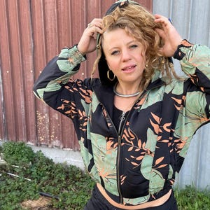 May include: A black hooded jacket with a floral print in shades of green and orange. The jacket has a zipper closure and a drawstring hood.