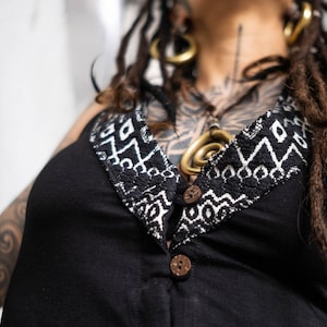 May include: Black sleeveless top with a decorative neckline featuring a white and black geometric pattern. The top has two wooden buttons and a V-neck design. The overall style suggests bohemian or alternative fashion.