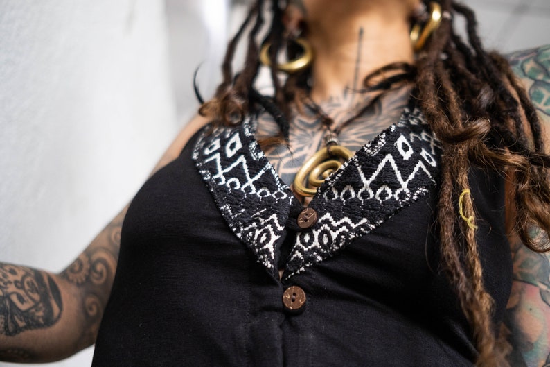 May include: Black sleeveless top with a decorative neckline featuring a white and black geometric pattern. The top has two wooden buttons and a V-neck design. The overall style suggests bohemian or alternative fashion.