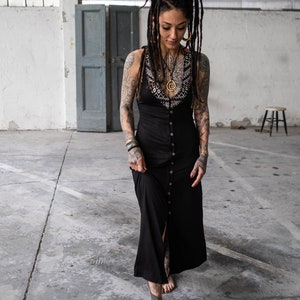 May include: A black maxi dress with a button-down front and a decorative neckline. The dress is worn by a person with tattoos and dreadlocks. The person is walking on a concrete floor.