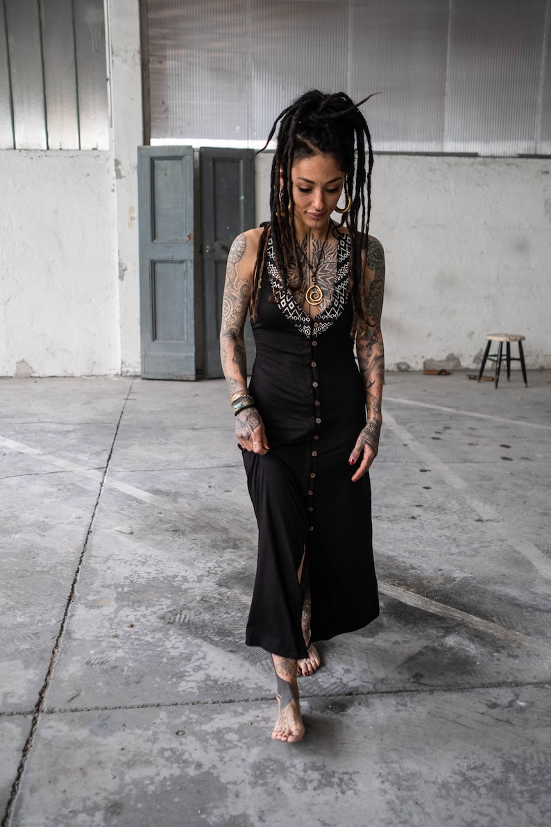 May include: A black maxi dress with a button-down front and a decorative neckline. The dress is worn by a person with tattoos and dreadlocks. The person is walking on a concrete floor.