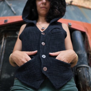 May include: Black hooded vest with large wooden buttons and two front pockets. The vest is made of a textured fabric. The person is wearing a necklace and gold bracelets. The background is an orange vehicle.