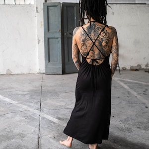 May include: A black maxi dress with a crisscross back detail. The dress is sleeveless and has a flowing skirt. The person wearing the dress has tattoos on their back and arms. The background is a concrete floor and a gray door.