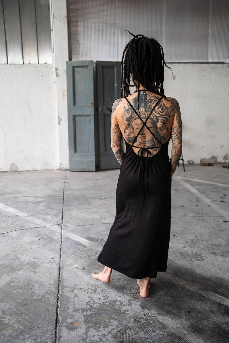 May include: A black maxi dress with a crisscross back detail. The dress is sleeveless and has a flowing skirt. The person wearing the dress has tattoos on their back and arms. The background is a concrete floor and a gray door.