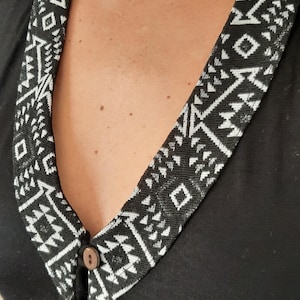 May include: Black sleeveless top with a decorative collar featuring a geometric black and white pattern. The collar is secured with three wooden buttons. The top is made of a soft, dark fabric.