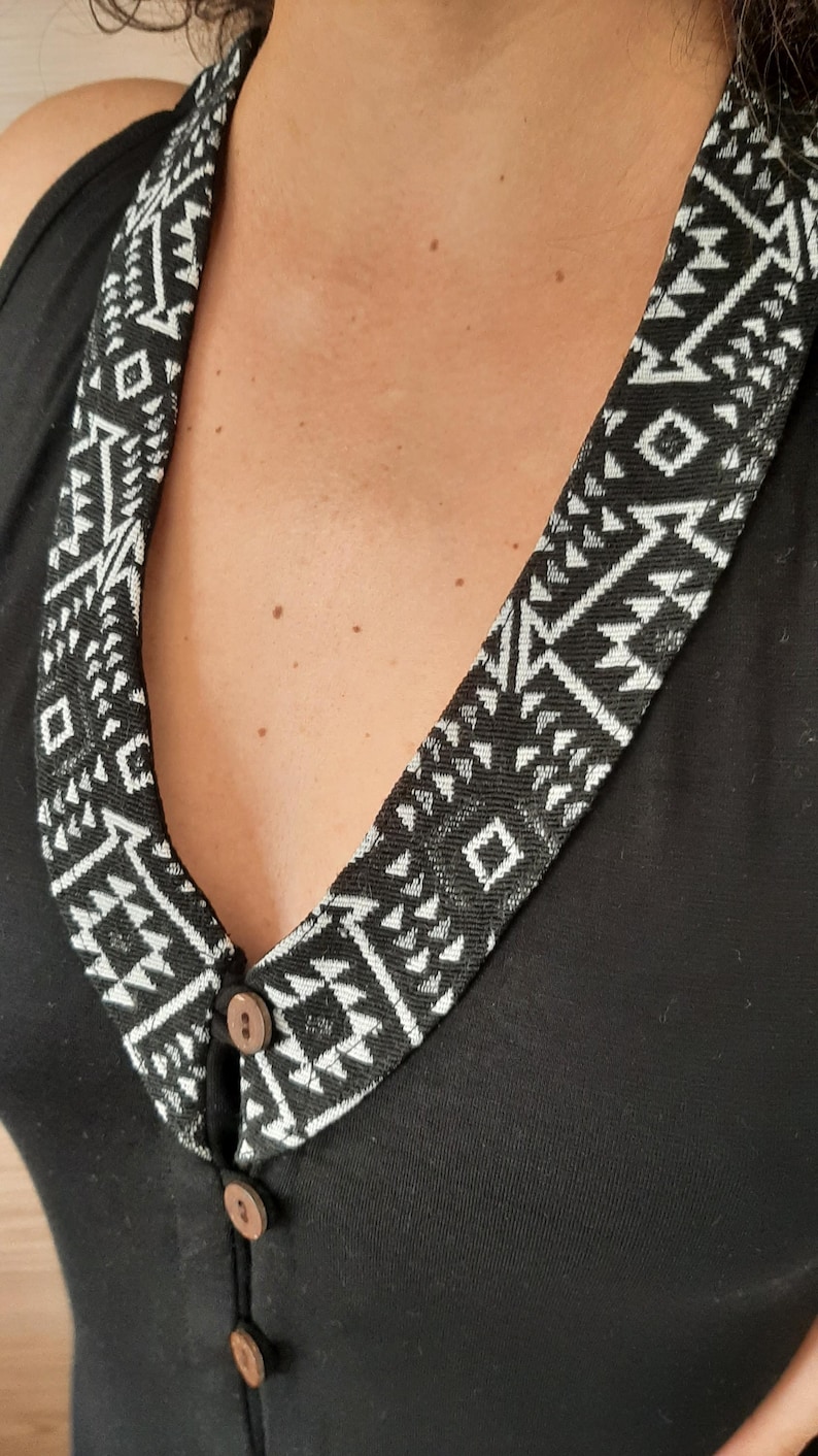 May include: Black sleeveless top with a decorative collar featuring a geometric black and white pattern. The collar is secured with three wooden buttons. The top is made of a soft, dark fabric.