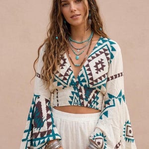 Peut inclure: Un haut court de couleur crème avec un décolleté en V profond, de longues manches évasées et une taille élastique smockée. Le vêtement est orné d'un motif géométrique dans des tons marron foncé et bleu sarcelle. Il est porté avec un pantalon blanc.