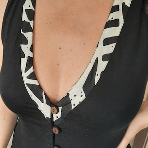 May include: Black sleeveless dress with a V-neckline and wooden buttons down the front. The neckline is trimmed with a cream-colored fabric featuring a black abstract pattern. The dress has a simple, elegant design.