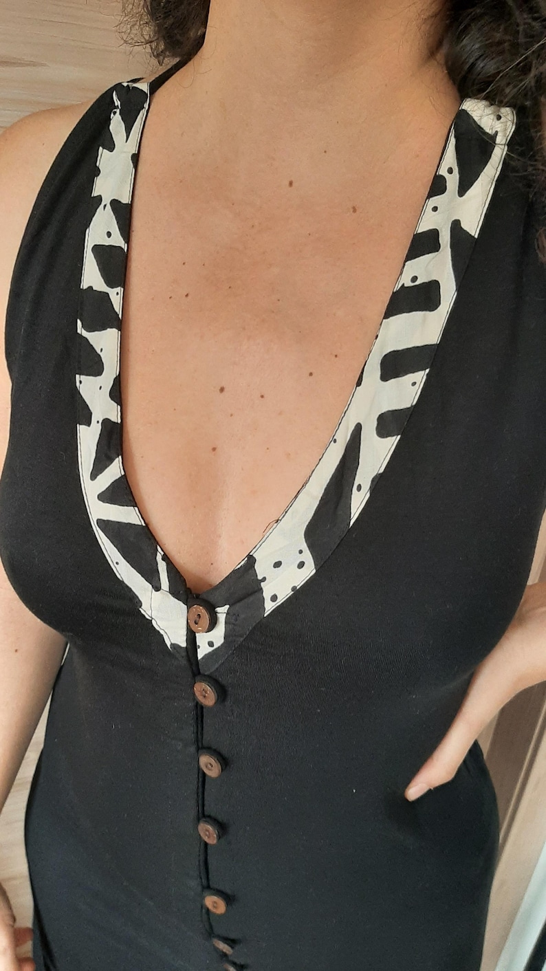 May include: Black sleeveless dress with a V-neckline and wooden buttons down the front. The neckline is trimmed with a cream-colored fabric featuring a black abstract pattern. The dress has a simple, elegant design.