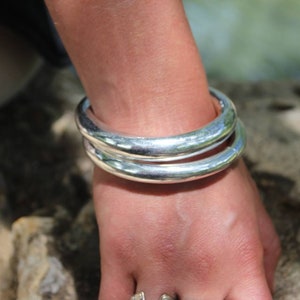 Op de afbeelding: Twee zilveren armbanden gestapeld op de pols van een persoon. De armbanden zijn glad en glanzend.