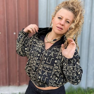 Puede incluir: Chaqueta negra con capucha y cremallera con un patrón geométrico crema. La chaqueta tiene una capucha y puños negros, y una cremallera color latón. La chaqueta se combina con pantalones negros. El estilo general es casual y bohemio.