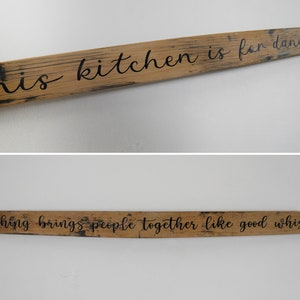May include: Two wooden signs with black lettering. The top sign reads "This kitchen is for dancing". The bottom sign reads "Nothing brings people together like good whisky".