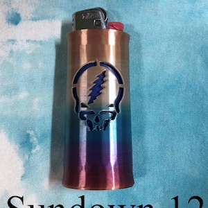 May include: A metal lighter with a gradient finish, transitioning from copper to blue and purple. It features a cut-out design of a skull with a lightning bolt. The text "Sundown 12" is at the bottom.