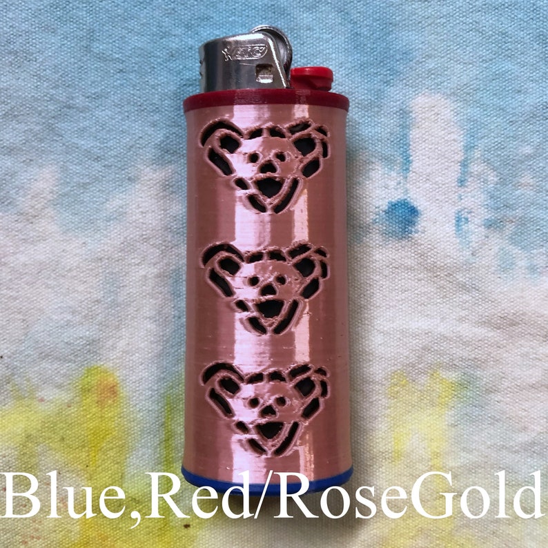 May include: A rose gold lighter with a blue and red top. The lighter has three cut-out bear faces on the side.