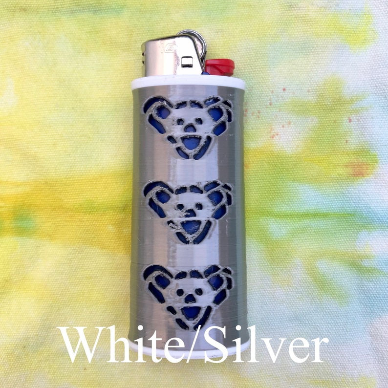 May include: A white and silver lighter with a design of three blue bear faces. The lighter is on a tie-dye background.