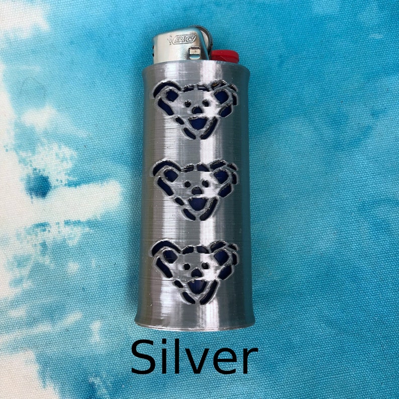 May include: A silver lighter case with three cut-out bear faces. The bear faces are outlined in blue. The lighter case is on a blue and white background. The word 'Silver' is written below the lighter case.