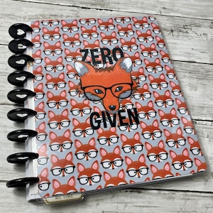 Può includere: Un'agenda con una copertina con un motivo ripetuto di volpi con gli occhiali su sfondo grigio. La copertina presenta anche una volpe con gli occhiali e le parole "ZERO GIVEN" in nero.